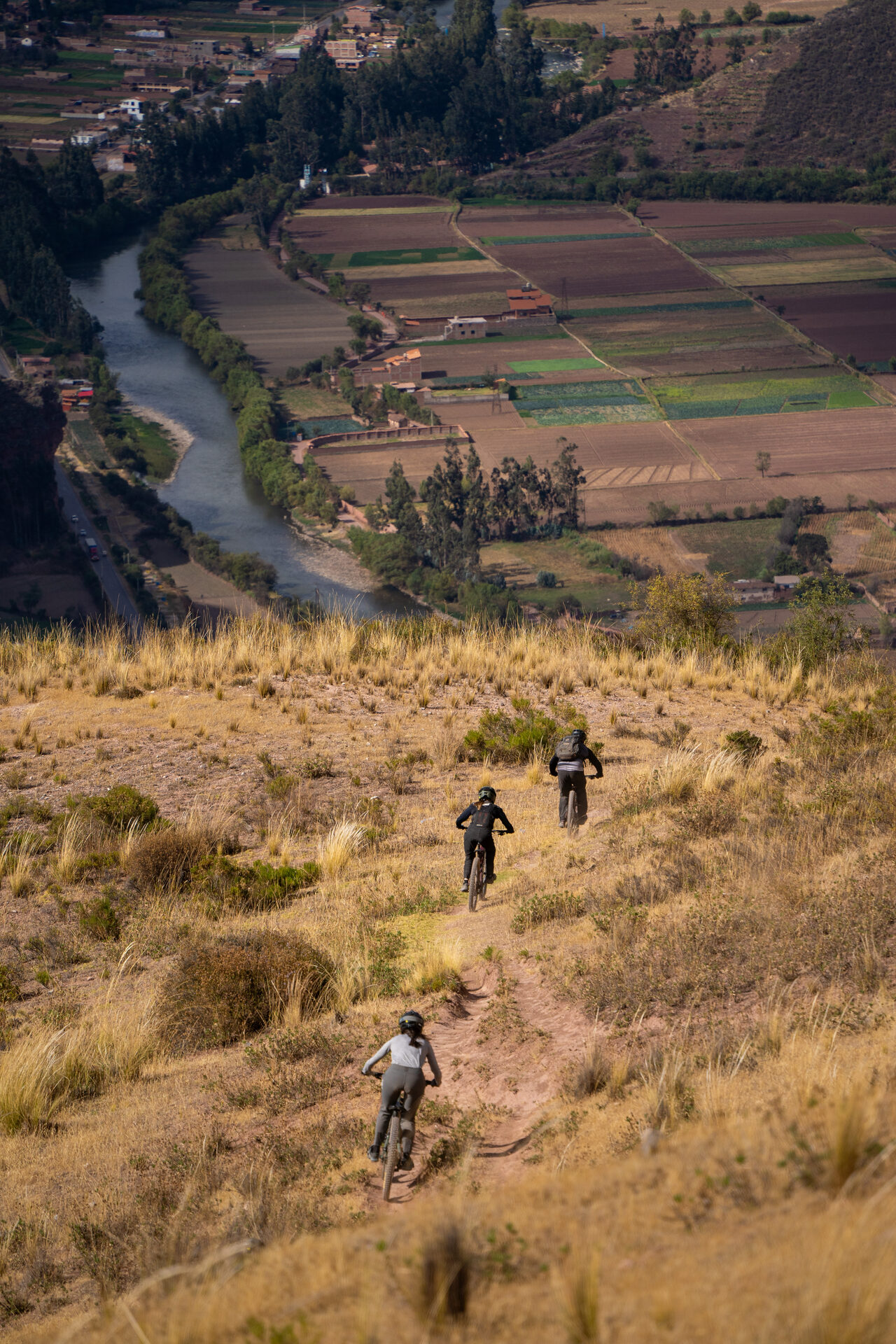 Three riders descending a ridge trail above the Sacred Valley river and terraced fields